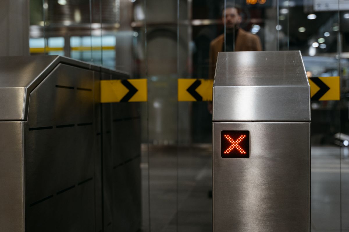 A stainless steel ticket barrier in a station, its red X lit to refuse entry, with a blurred figure behind it