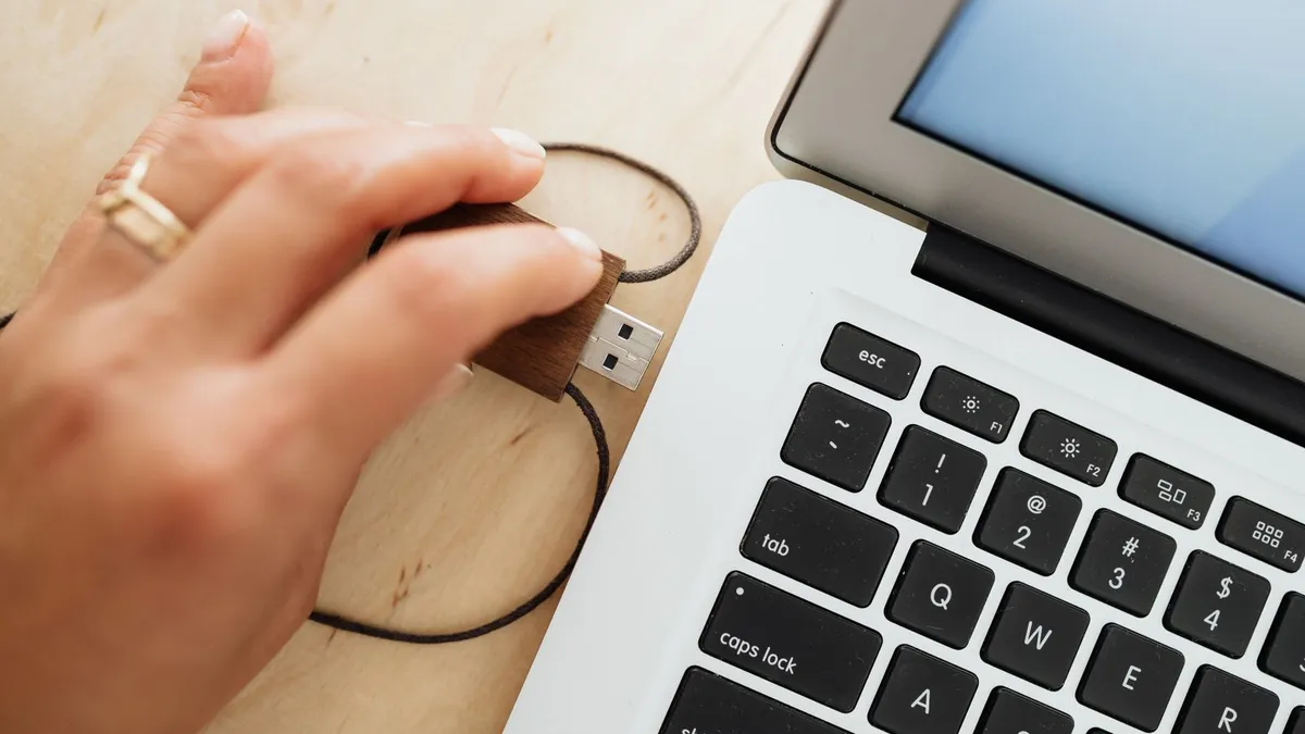 A hand wearing a gold ring plugs a wooden USB stick on a fabric cord into the USB port of a white laptop, on a light wooden desk
