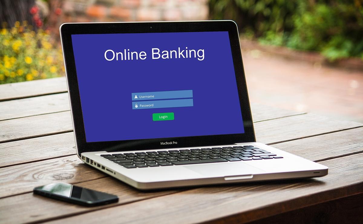 A laptop showing an online banking login screen with username and password fields, a phone beside it on a wooden table — phishing pages imitate real logins like this to capture your credentials.